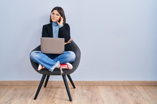 Young Hispanic Woman Sitting On Chair Using Computer Laptop Looking Confident At The Camera Smiling With Crossed Arms And Hand Raised On Chin. Thinking Positive.