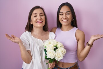 Hispanic mother and daughter holding bouquet of white flowers smiling cheerful presenting and pointing with palm of hand looking at the camera.