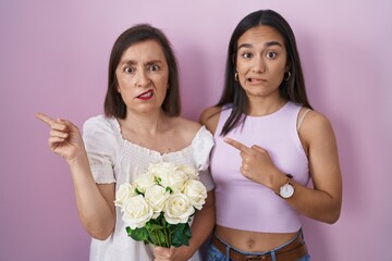 Hispanic mother and daughter holding bouquet of white flowers pointing aside worried and nervous...
