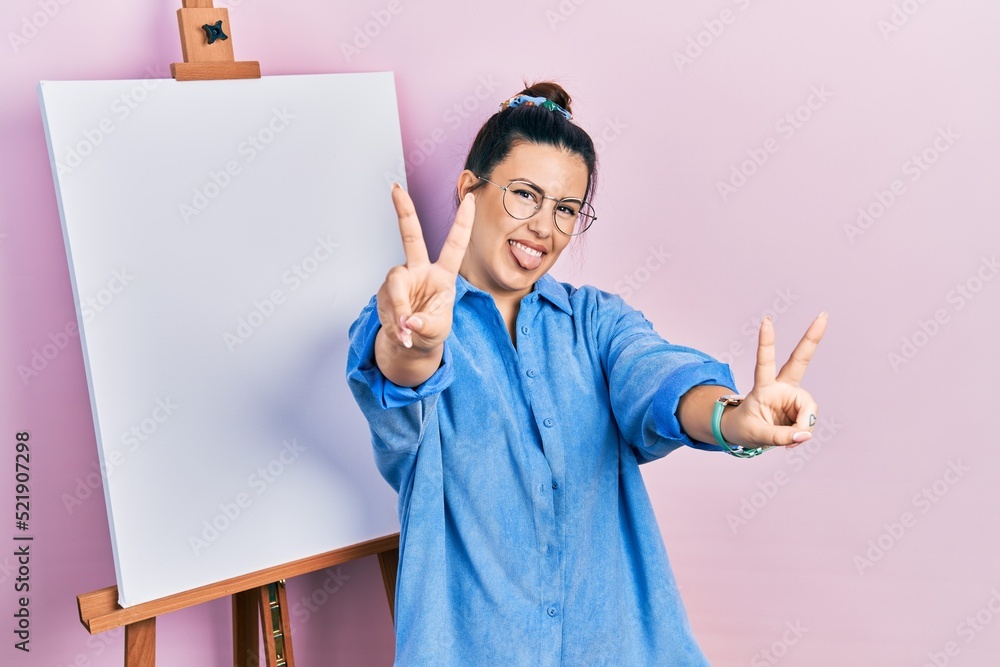 Poster young hispanic woman standing by painter easel stand smiling with tongue out showing fingers of both