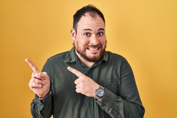 Plus size hispanic man with beard standing over yellow background pointing aside worried and...