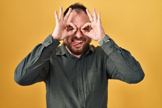 Plus Size Hispanic Man With Beard Standing Over Yellow Background Doing Ok Gesture Like Binoculars Sticking Tongue Out, Eyes Looking Through Fingers. Crazy Expression.