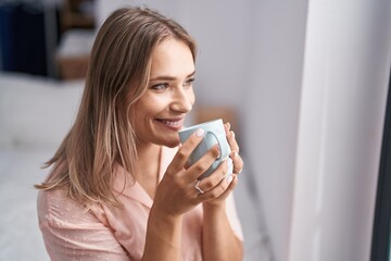 Young caucasian woman drinking cup of coffee sitting on bed at bedroom