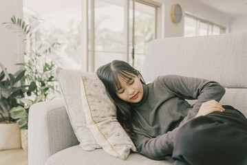 Young Asian woman lying down on sofa at home, stomach pain, gut health care concept