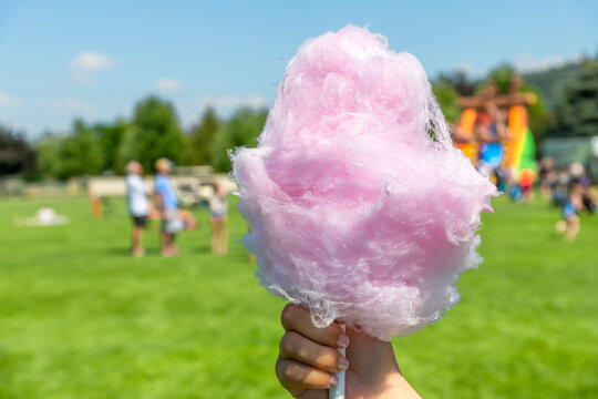 A hand holding pink cotton candy at an outdoor summer fair with festival goers blurred behind.