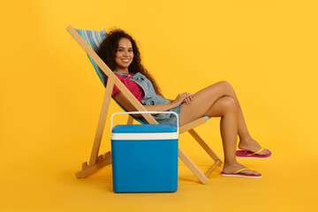 Happy young African American woman resting in deck chair near cool box on yellow background