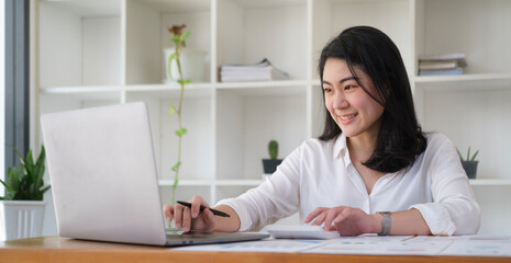 Asian businesswoman working with laptop and using calculator at her office desk.