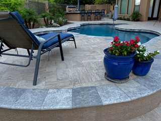 A travertine tiled pool deck with a spa and outdoor kitchen on a desert landscaped backyard in...