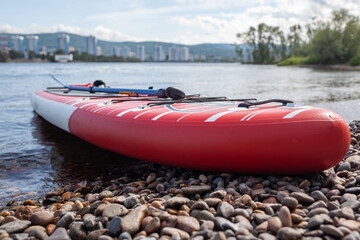 Red SUP board on the river bank. Paddle board on the lake shore. Supboard water tourism and active sports.