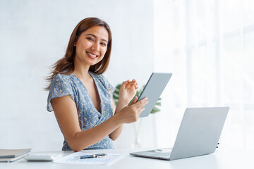 Looking camera, Young pretty Asian business woman working with laptop computer and tablet pc in modern office desk workplace.