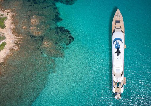 White big yacht for a billionaire anchored off a rocky shore aerial view. Mega yacht on blue water aerial view. Modern big white yacht on transparent water, coastline top view.