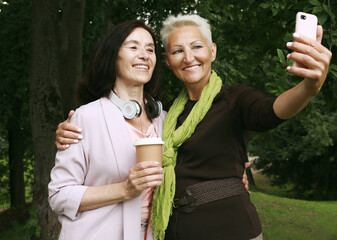 Two charming elderly women friends holds a cup of coffee to go and making selfie in summer park.