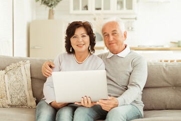 Pretty elderly 60s couple resting on couch in living room hold on lap laptop watching movie smiling enjoy free time