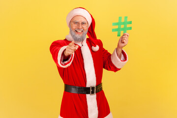 Positive elderly man with gray beard wearing santa claus costume standing with green hashtag in hands, pointing to camera with toothy smile. Indoor studio shot isolated on yellow background.