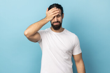 Portrait of unlucky man with beard wearing white T-shirt standing with facepalm gesture, feeling regret and sorrow, blaming herself for mistake. Indoor studio shot isolated on blue background.