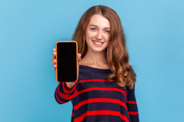 Smiling woman wearing striped casual style sweater, showing smart phone with empty display for advertisement, looking at camera with happy face. Indoor studio shot isolated on blue background.
