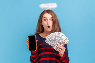 Shocked woman wearing striped casual style sweater and nimb over head, shows cell phone with empty display and dollars, has astonished expression. Indoor studio shot isolated on blue background.