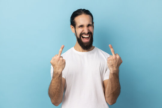 Portrait of man with beard wearing white T-shirt showing middle fingers, rude gesture, aggressive protest with impolite finger sign, expressing hatred. Indoor studio shot isolated on blue background.