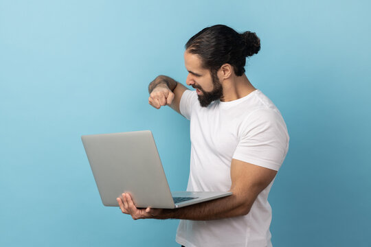 Portrait of aggressive man with beard wearing white T-shirt working on laptop computer, has mistake, wants to bang her fist on the screen. Indoor studio shot isolated on blue background.