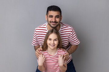 Portrait of delighted smiling father and daughter in striped T-shirts standing and looking at camera, dad embracing little kid, expressing happiness. Indoor studio shot isolated on gray background. - Powered by Adobe