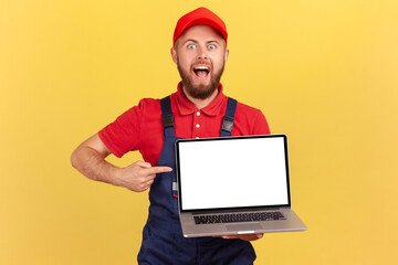 Portrait of excited bearded handyman in overall holding laptop with empty display, pointing at screen and looking at camera with open mouth. Indoor studio shot isolated on yellow background.