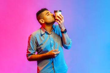 Portrait of handsome young adult man in shirt kissing disposable cup of take away coffee, enjoying...