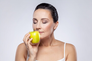 Obraz na płótnie Canvas Closeup portrait of beautiful charming middle aged woman holding smelling green apple, vitamins for beauty and skin care, keeps eyes closed. Indoor studio shot isolated on gray background.
