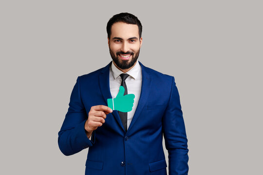 Portrait of happy handsome bearded businessman looking at camera with smile and showing paper thumb up, wearing official style suit. Indoor studio shot isolated on gray background.