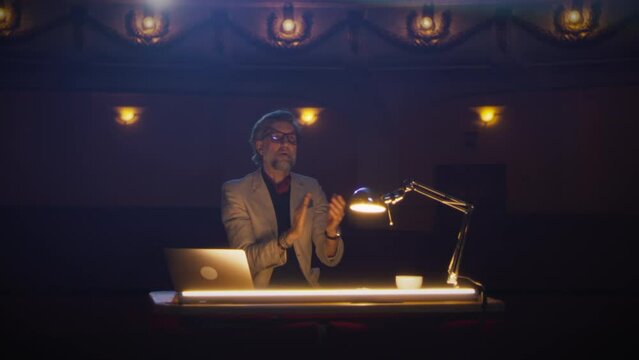 During the rehearsal of the performance, male director in a suit rejoicing and clapping his hands to the actors in the dim theater