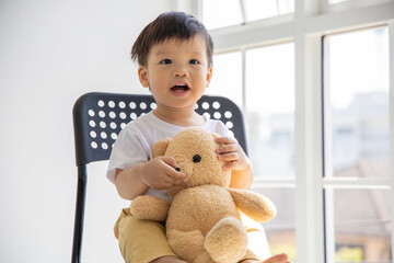 Little asian boy sitting on black chair and holding bear doll with smile in front of big window at home.