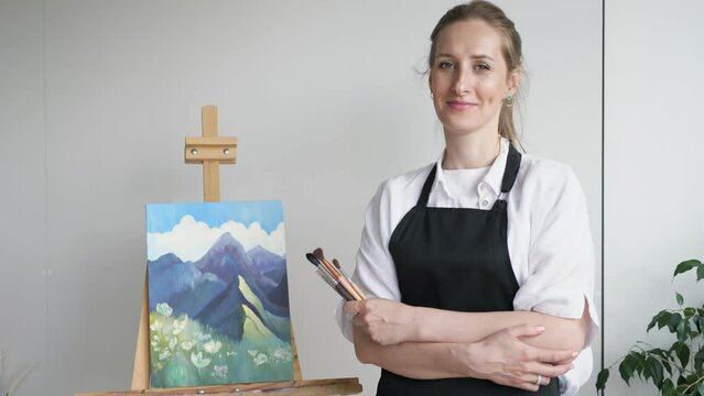 Brown-haired woman smiles and shows enjoyment of favourite hobby. Mature housewife with crossed arms stands against created watercolor picture of landscape closeup