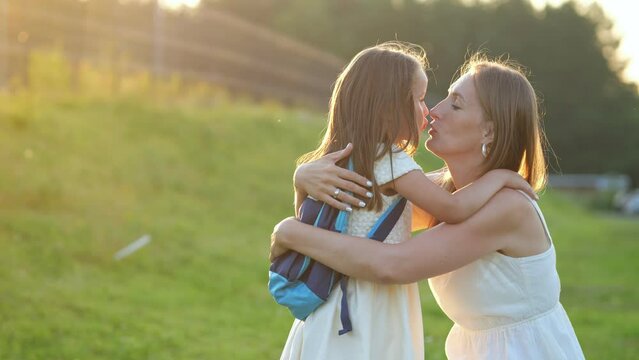 Mother meets happy first-grader daughter and kisses on cheeks standing near school. Smiling parent interested about first day at elementary school at sunset