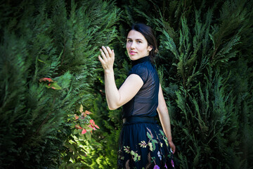 Naklejka premium A young pretty woman in black clothes is standing near a bush. Photography in a dark key