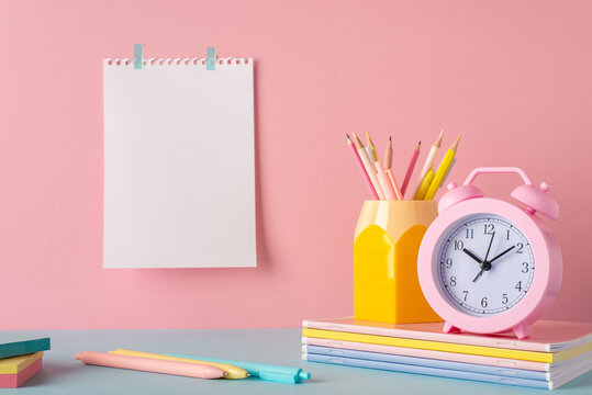 Back to school concept. Photo of school accessories on blue desk pencil holder alarm clock over stack of notebooks pens and paper sheet attached to pink wall with empty space