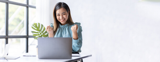 Happy excited successful Asian businesswoman triumphing with a laptop computer in the workplace...