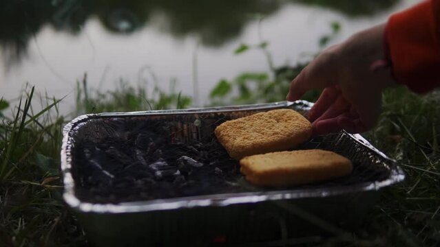 Video of two breads being cooked next to a water body on a cast-iron BBQ grill using charcoal.