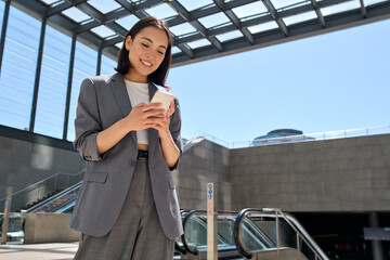 Young smiling Asian business woman wearing suit holding cell phone standing in city metro using...