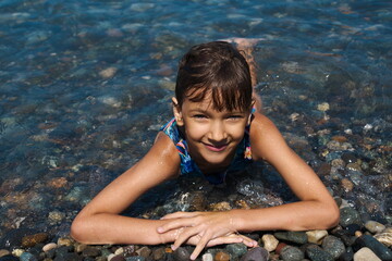 kid female of 7 years old lies in the shallow water on the  stones in sea, wet happy smiling looks up at the camera, sunny