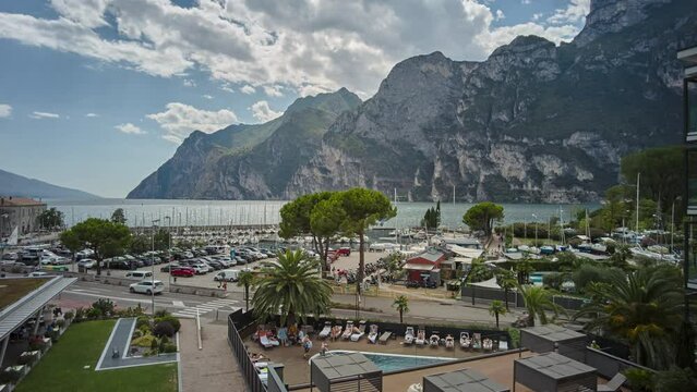 Time lapse of the port area of Riva del Garda and the clouds moving over the lake and the mountains. Transport of vehicles along the lakefront and tourists who enjoy the services of the place.