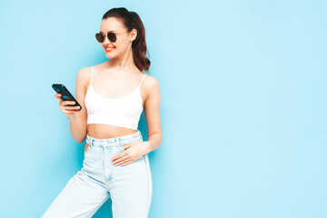 Fototapeta na wymiar Young beautiful smiling female in trendy summer jeans. Sexy carefree woman posing near blue wall in studio. Positive brunette model looking at cellphone screen. Holding smartphone and using apps