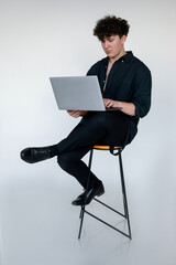 Full length photo of upset young curly guy in total black look sitting cross legged on chair and playing computer games on modern laptop, gray studio background. Modern Internet reality