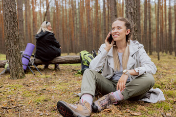 Mom and daughter in the forest. Woman with blonde hair tied in braids sits on the ground in the forest and talks on the phone, resting while trekking, camping, hiking.