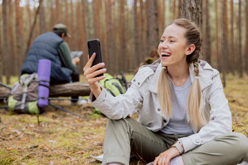 Smiling woman with blonde hair tied in braids sits on the ground in the forest and talks on the phone video conversation, resting while trekking, camping, hiking.