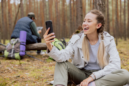 Smiling woman with blonde hair tied in braids sits on the ground in the forest and talks on the phone video conversation, resting while trekking, camping, hiking.