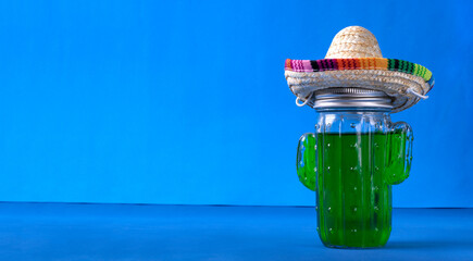Green drink in cactus shaped glass jar with sombrero hat on blue background. Refreshing summer beverage. Mexican vacation concept. Mockup with copy space - 521951444