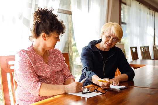 Portrait of two smiling elderly women sitting at brown desk, talking, discussing, making notes in notepad in large room.