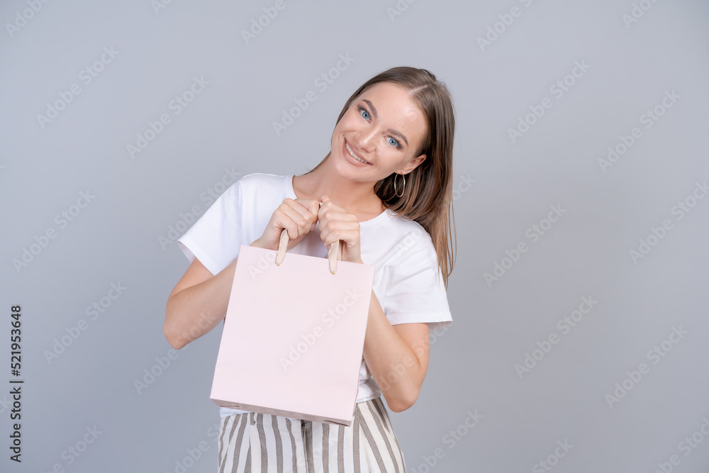 Wall mural Teen girl doing shopping holding shopping bag in her hands fashion young woman shopper isolated on gray background. Beautiful happy girl in white light clothes. Copy space and mock up. Seasonal sales