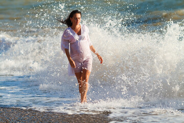 Pregnant woman is jumping on beach. Cheerful pregnant woman runs on seashore. Pregnant woman in a romantic white dress jumping on the beach near the tropical sea. Indication of happy pregnant women.