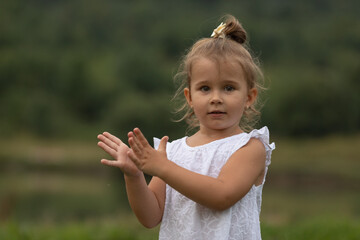 A cute little girl in a white dress stands on the shore of a lake on a summer evening and claps her hands. Sunset on the lake.