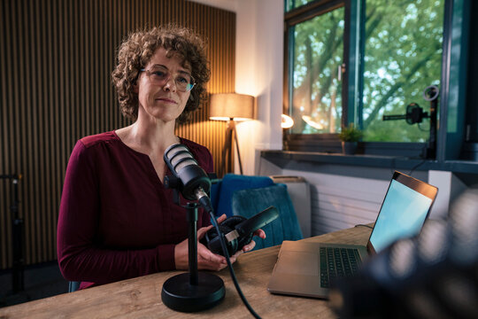Mature presenter wearing eyeglasses holding headset sitting with laptop at desk in radio station